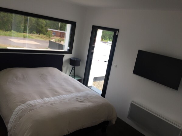 A bedroom with a bed, a desk, a chair, a lamp, and a window with a view of a building and greenery.