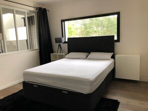 A modern bedroom with a bed, a nightstand, a lamp, and a view of trees through a window. - Modern house on La Baule international golf course (Saint-André-Des-Eaux)