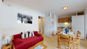 A compact living space with a red sofa, a dining table set for four, a kitchen area, and a wall-mounted picture of a snowy landscape. - Balcons B 026 - PARC NAT. VANOISE appart. 6 pers. (Val Cenis)