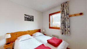 A bedroom with a wooden headboard, two white beds, a red blanket, a nightstand with a lamp, a window with curtains, and a wall-mounted heater. - Balcons B 026 - PARC NAT. VANOISE appart. 6 pers. (Val Cenis)