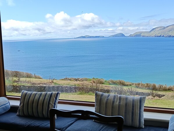 A view from a window overlooking a large body of water with distant landforms, a stone wall, and a seating area with striped cushions.