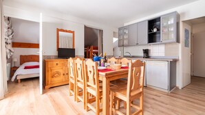 A compact room with a wooden dining table, chairs, a small kitchenette, a bed, and a television.