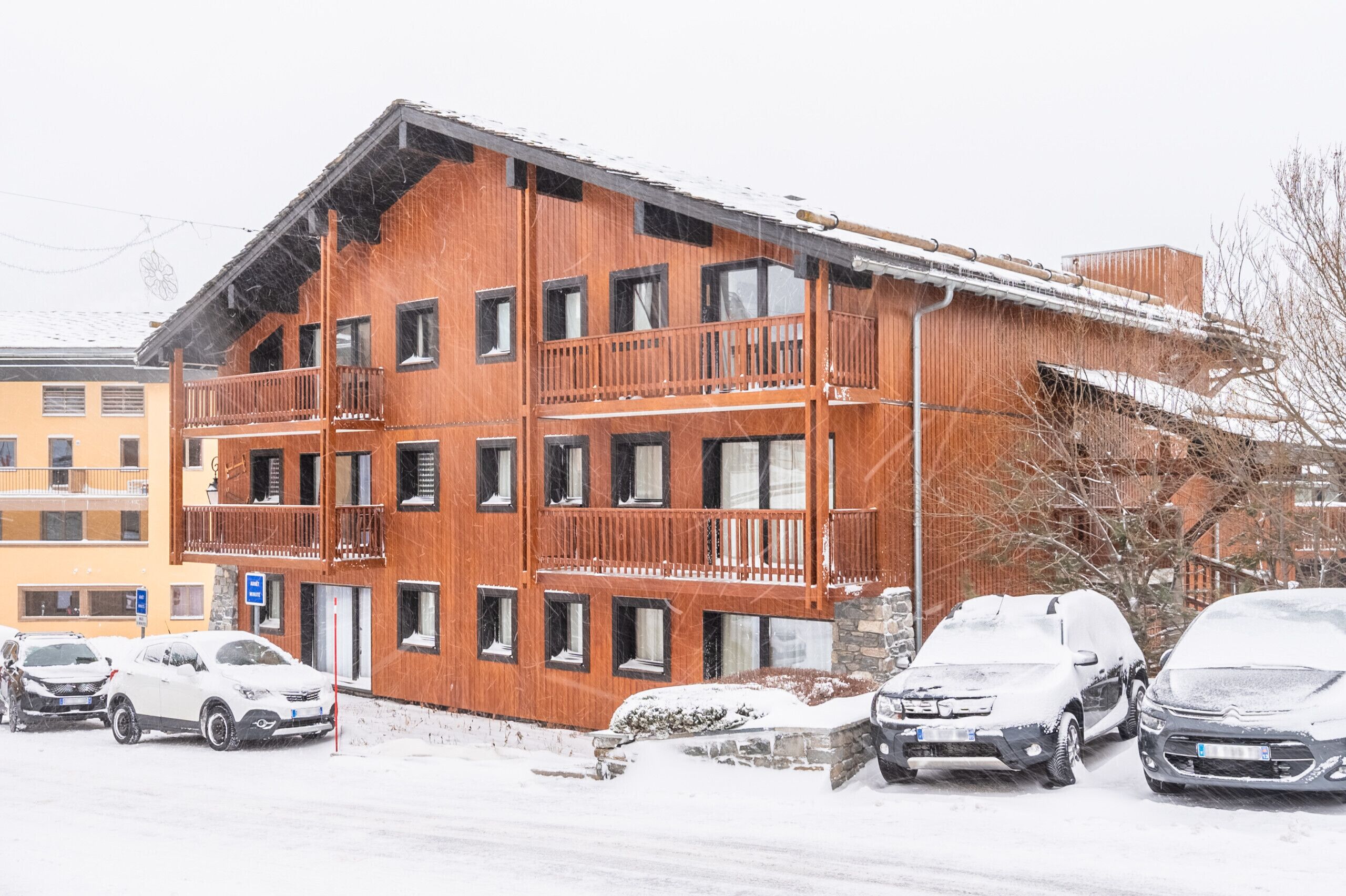 Ein mehrstöckiges Holzgebäude mit Balkonen, einem Parkplatz mit Autos und einer verschneiten Umgebung.