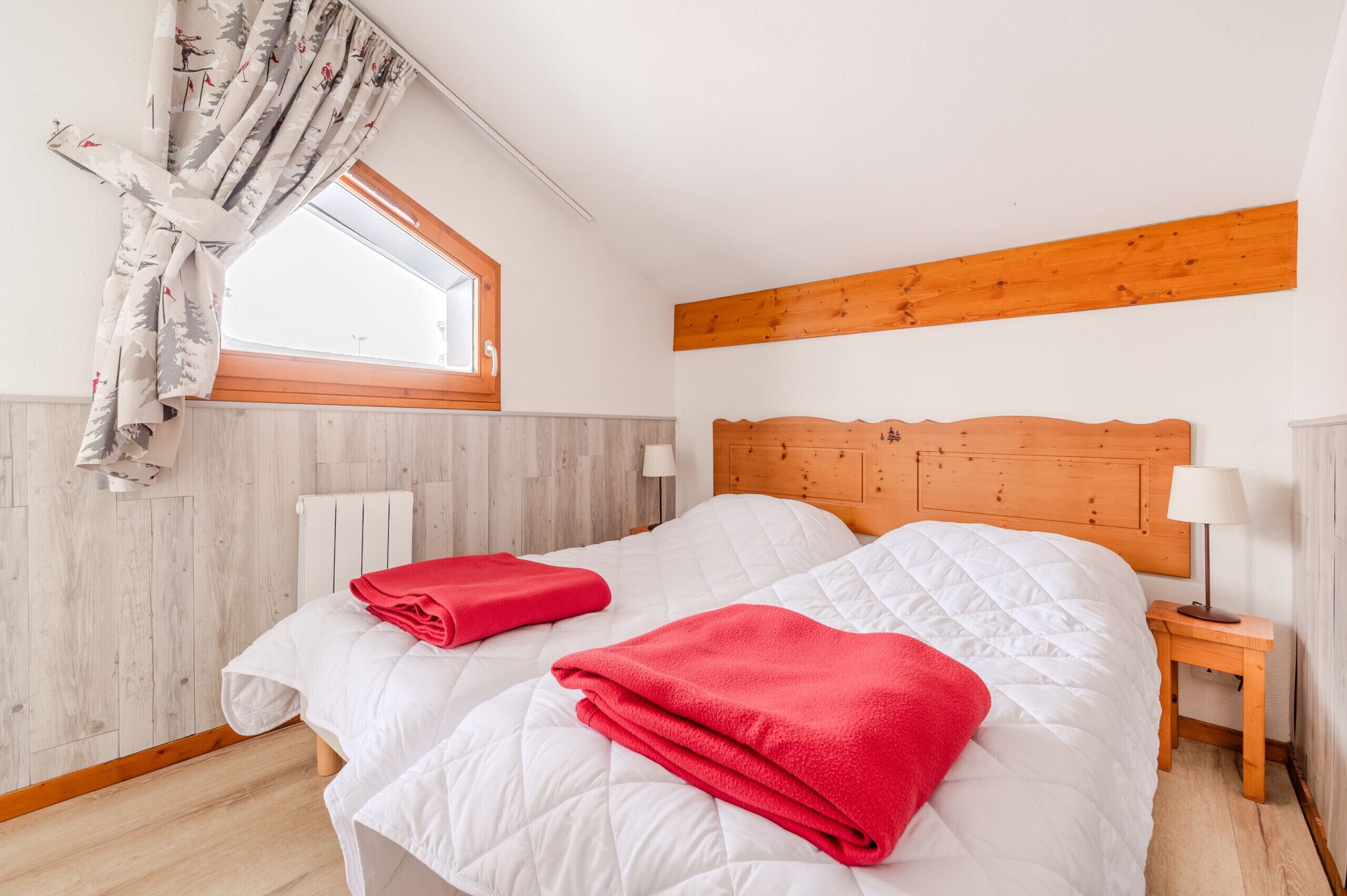 Un dormitorio con dos camas, cabecero de madera, una ventana con cortinas, una mesita de noche con lámpara y un radiador.