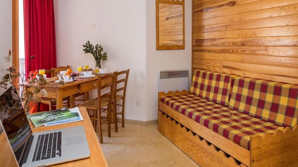 Ein gemütliches Zimmer mit einer Holzwand, einem karierten Sofa, einem Holzesstisch mit Stühlen und einem Laptop auf dem Tisch.