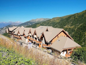 A row of wooden chalets with sloped roofs, set against a mountainous backdrop.