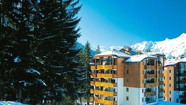 Een hotel met meerdere verdiepingen en balkons, omgeven door besneeuwde bergen en bomen.