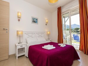 Ein Hotelzimmer mit einem Bett, zwei Nachttischlampen, einem Nachttisch, einem Fenster mit Vorhängen und Blick ins Freie.