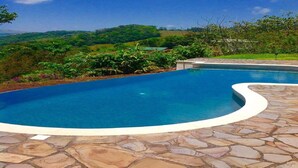 Une piscine dotée d’une plage en pierre, entourée d’une végétation luxuriante et avec des collines en arrière-plan.