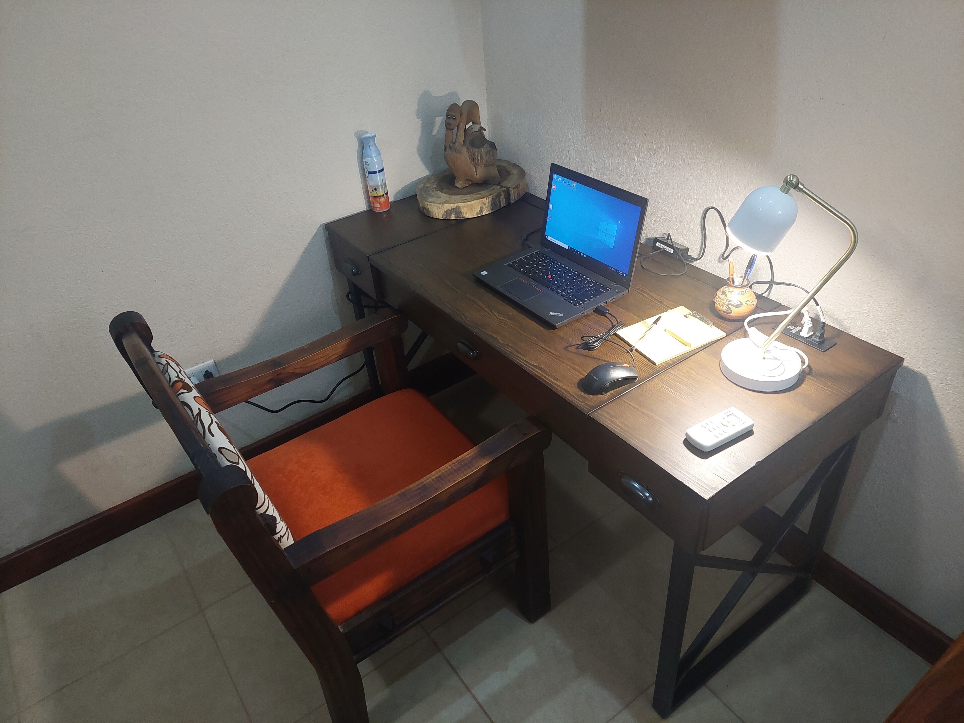Un escritorio de madera con una computadora portátil, un ratón, una lámpara y una silla con un cojín naranja.