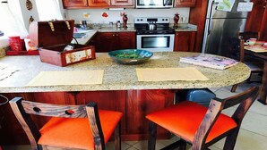 Une cuisine avec des meubles de rangement en bois, un plan de travail en granit, une cuisinière et deux chaises orange.