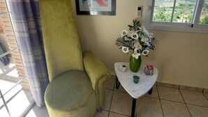 A room with a green armchair, a framed abstract painting, a small table with a vase of flowers, and a window with a view of greenery and hills.