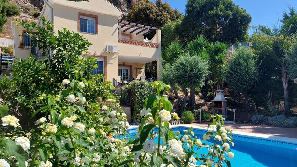 A poolside view of a building with a pergola, surrounded by lush greenery and a clear blue sky.