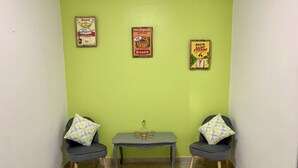 Um quarto pequeno com duas poltronas cinzentas, uma mesinha com uma garrafa e copos, e três cartazes emoldurados na parede.