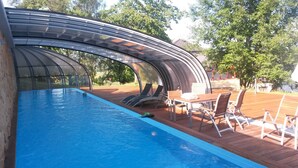 Una piscina con cerramiento de vidrio curvo, rodeada por una terraza de madera y mobiliario exterior.