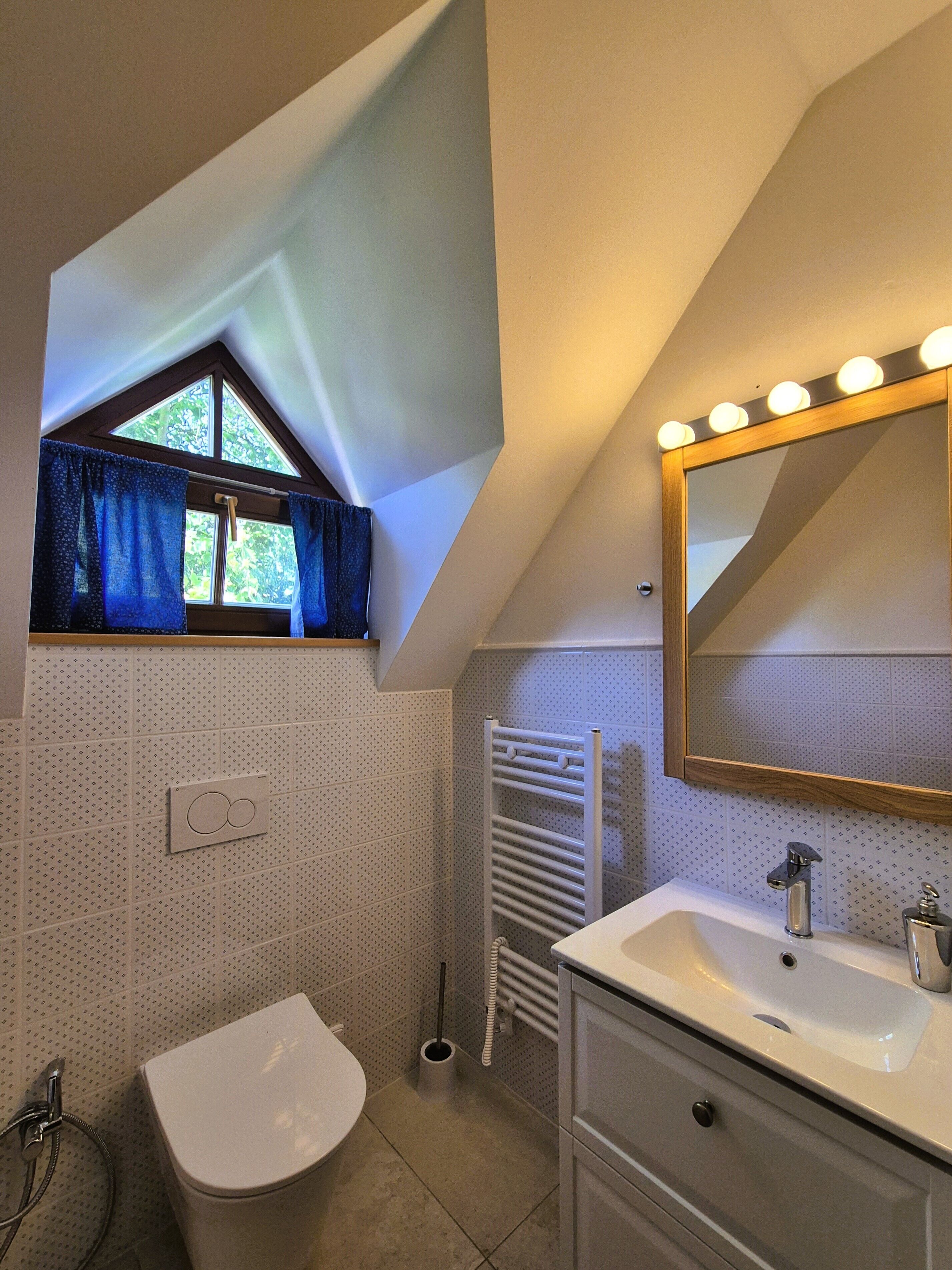 A bathroom with a white sink, a mirror with lights, a toilet, a window with curtains, and a towel rack.