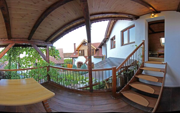 A wooden balcony with a curved roof, a wooden bench, a staircase with a handrail, and a view of houses in the background.