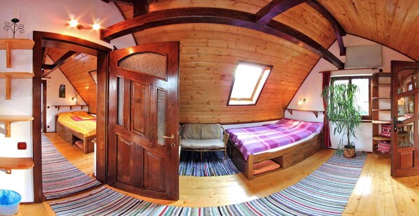 Un dormitorio acogedor en el ático, con techo inclinado, una puerta de madera grande, una cama con manta morada, un sofá pequeño y una planta en maceta.