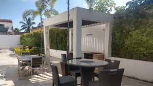 Eine Terrasse im Freien mit einem runden Tisch, Stühlen, einer Pergola und umliegendem Grün.