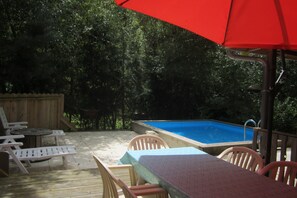 A wooden deck with a pool, outdoor furniture, and a red umbrella. - Wooden lodge with terrace and swimming pool in a green setting (Les Philiberts)