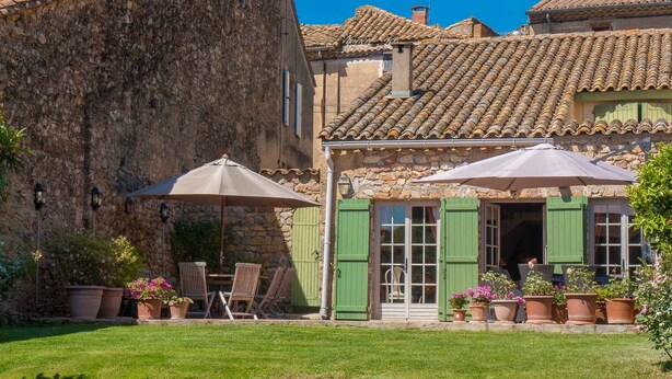 A stone building with a tiled roof, a grassy courtyard, and outdoor seating with umbrellas.