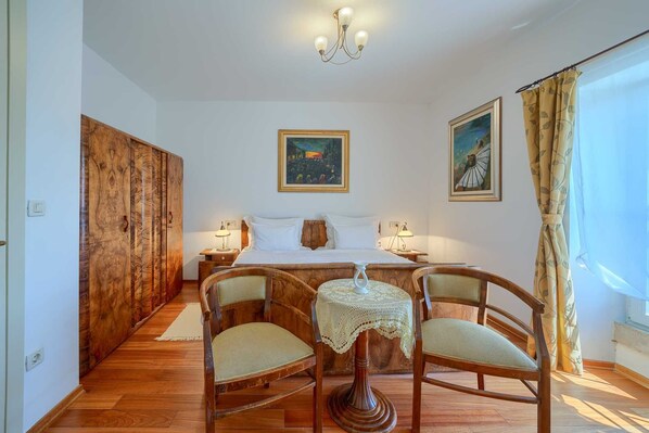 A bedroom with a wooden wardrobe, a bed with white linens, two bedside tables with lamps, a wooden table with a lace tablecloth, and two chairs.