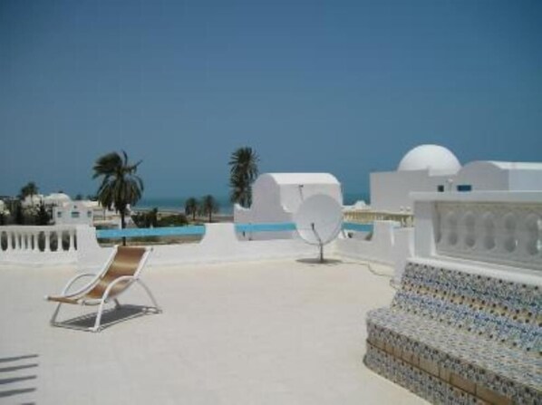 Une terrasse sur le toit, avec un fauteuil de relaxation, un canapé  à motifs et une vue sur la mer et les palmiers.