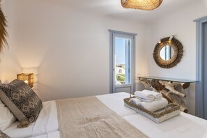 A bedroom with a bed, a bedside lamp, a window, a mirror, and a wall-mounted shelf with towels.