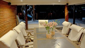 A covered outdoor dining area with a table and chairs, a view of the beach, and a wooden wall.