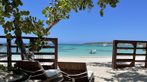 Two wooden lounge chairs facing a beach with clear blue water and boats.