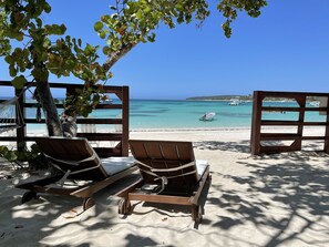 Two wooden lounge chairs facing a beach with clear blue water and boats.