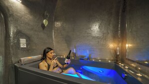 Une personne dans un bain à remous de couleur sombre, éclairé de lumières bleues, tenant une bouteille et un verre.