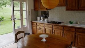 A kitchen with wooden cabinets, a white countertop, and a dining table with a white cake stand. - Small house o waterfront. (Valdallière)