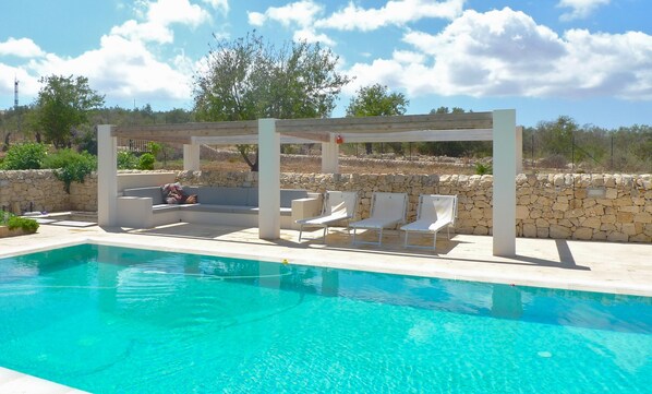A pool area with a pergola, lounge chairs, and a stone wall.