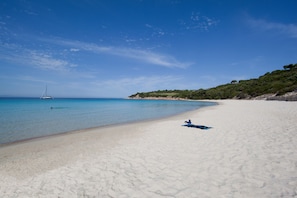 Une plage de sable blanc, un voilier sur l’eau et une serviette bleue sur le sable.