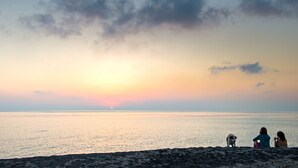 A sunset over a calm sea with two people and a dog on the shore.