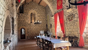 Una habitación con paredes de piedra y un techo alto en arco, una larga mesa dispuesta para comer y una estatua de un ángel.