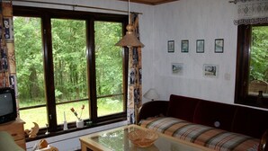 Un salon chaleureux avec un canapé, un fauteuil et une table basse. De grandes fenêtres offrent une vue sur la verdure environnante.