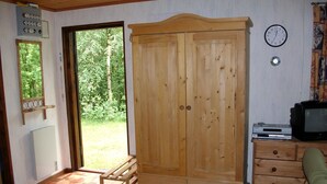 Une pièce avec une armoire en bois, un téléviseur posé sur un meuble en bois, une horloge au mur et une fenêtre donnant sur un paysage verdoyant.