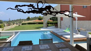 A poolside area with a wooden pergola, lounge chairs, and a stone wall. - Pefkochori villa with a stunning panoramic view and a waterfall private pool. (Pefkochori Chalkidiki)