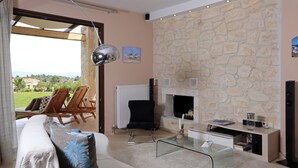 A living room with a stone wall, a fireplace, a glass coffee table, and a sofa with decorative pillows. - Pefkochori villa with a stunning panoramic view and a waterfall private pool. (Pefkochori Chalkidiki)