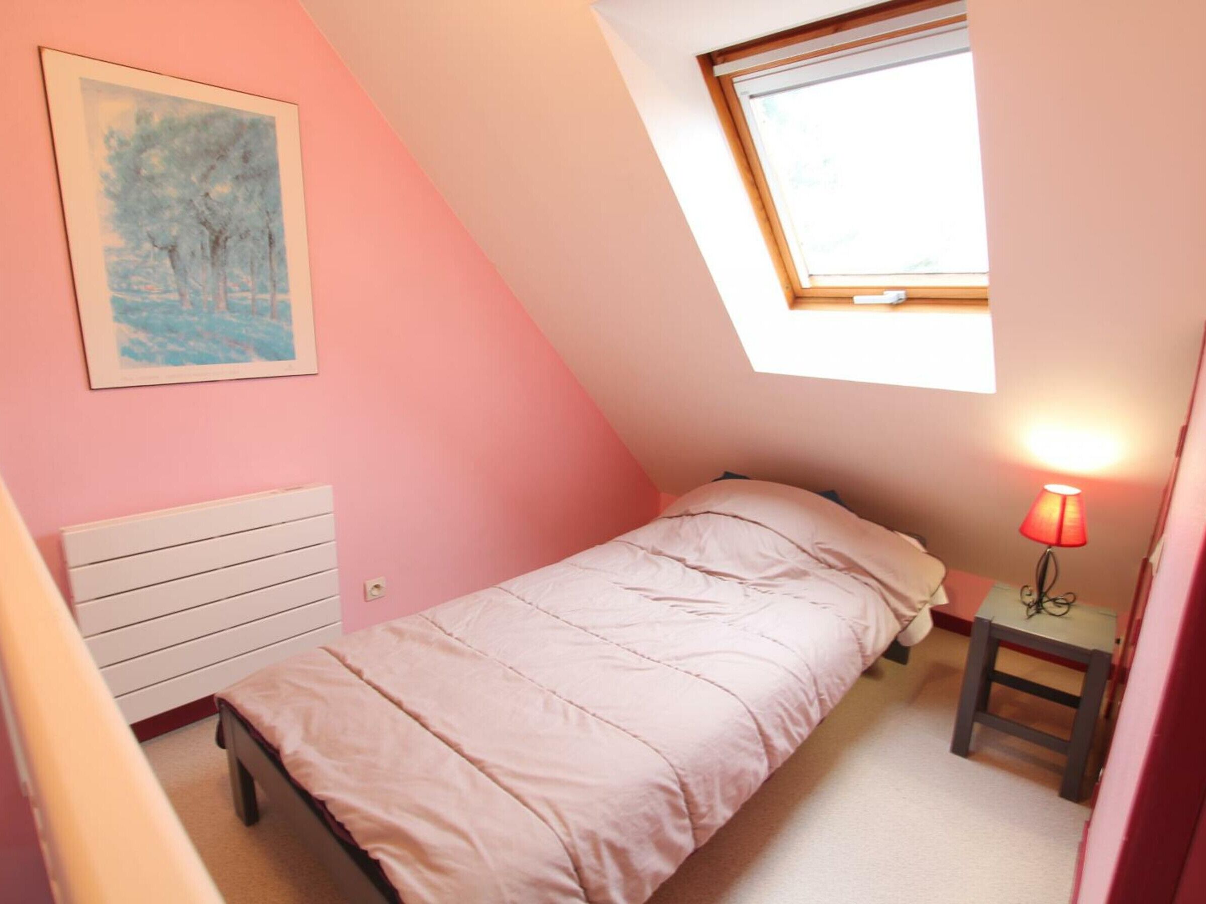 Una habitación pequeña con cama individual, techo inclinado, ventana, mesita de noche con lámpara y radiador.