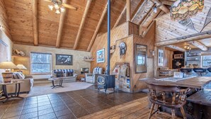 Interior rústico de cabana com teto alto, vigas de madeira, área de jantar com mesa e cadeiras de madeira, cozinha com micro-ondas e um espaço de estar acolhedor com sofá e poltronas.
