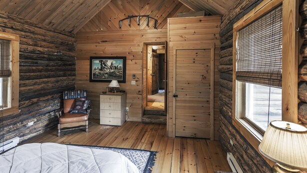 Quarto de cabana de toras com uma cama, uma cadeira, um aparador, um abajur e um quadro na parede.