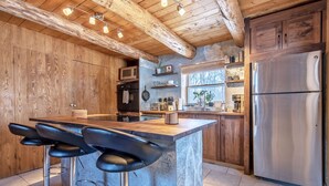 Uma cozinha moderna com ilha central, banquetas, armários de madeira, uma geladeira de aço inoxidável e uma janela que traz luz natural.