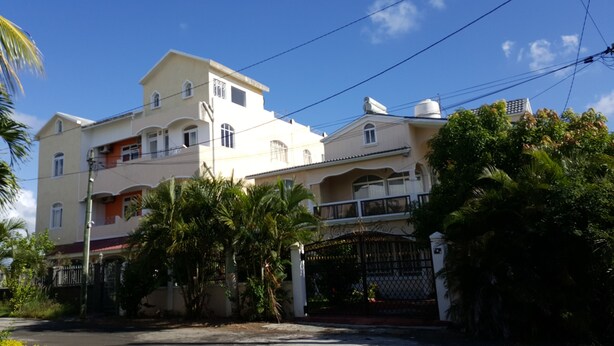 Un edificio residencial de varios pisos con balcones, entrada protegida por un portón y vegetación circundante.