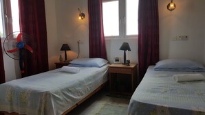 Una habitación con dos camas individuales, una mesita de noche con lámpara, una pequeña mesa con lámpara, un calentador en la pared y una ventana con cortinas.