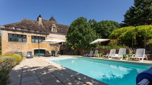 Una piscina con sillones de descanso y sombrillas, un edificio de piedra con torreón y exuberante vegetación.