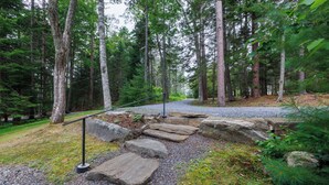 A forested area with a gravel path, wooden steps, and a metal railing. - Water's Edge - Waterfront Cottage on Southport - Wrap-Around Porch! (Boathouse Lane)