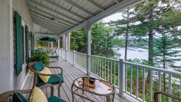 A covered porch with wicker furniture and a glass table, overlooking a forest and a body of water. - Water's Edge - Waterfront Cottage on Southport - Wrap-Around Porch! (Boathouse Lane)